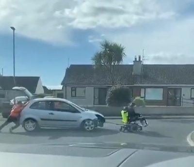 Dos personas tratan de remolcar un coche con una silla de ruedas motorizada.